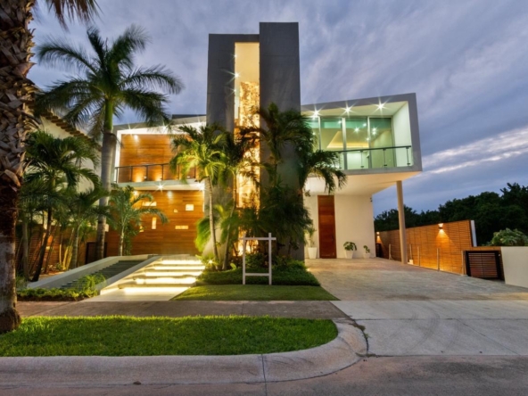 Fachada iluminada de una casa de lujo en venta en Puerto Cancún, con diseño moderno, palmeras y entrada principal elevada con escalinata.