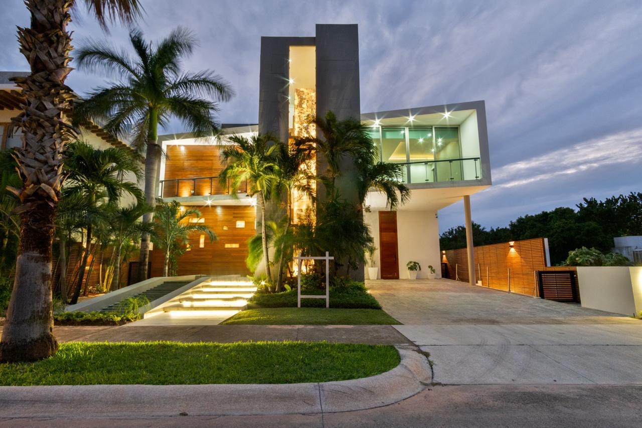 Fachada iluminada de una casa de lujo en venta en Puerto Cancún, con diseño moderno, palmeras y entrada principal elevada con escalinata.